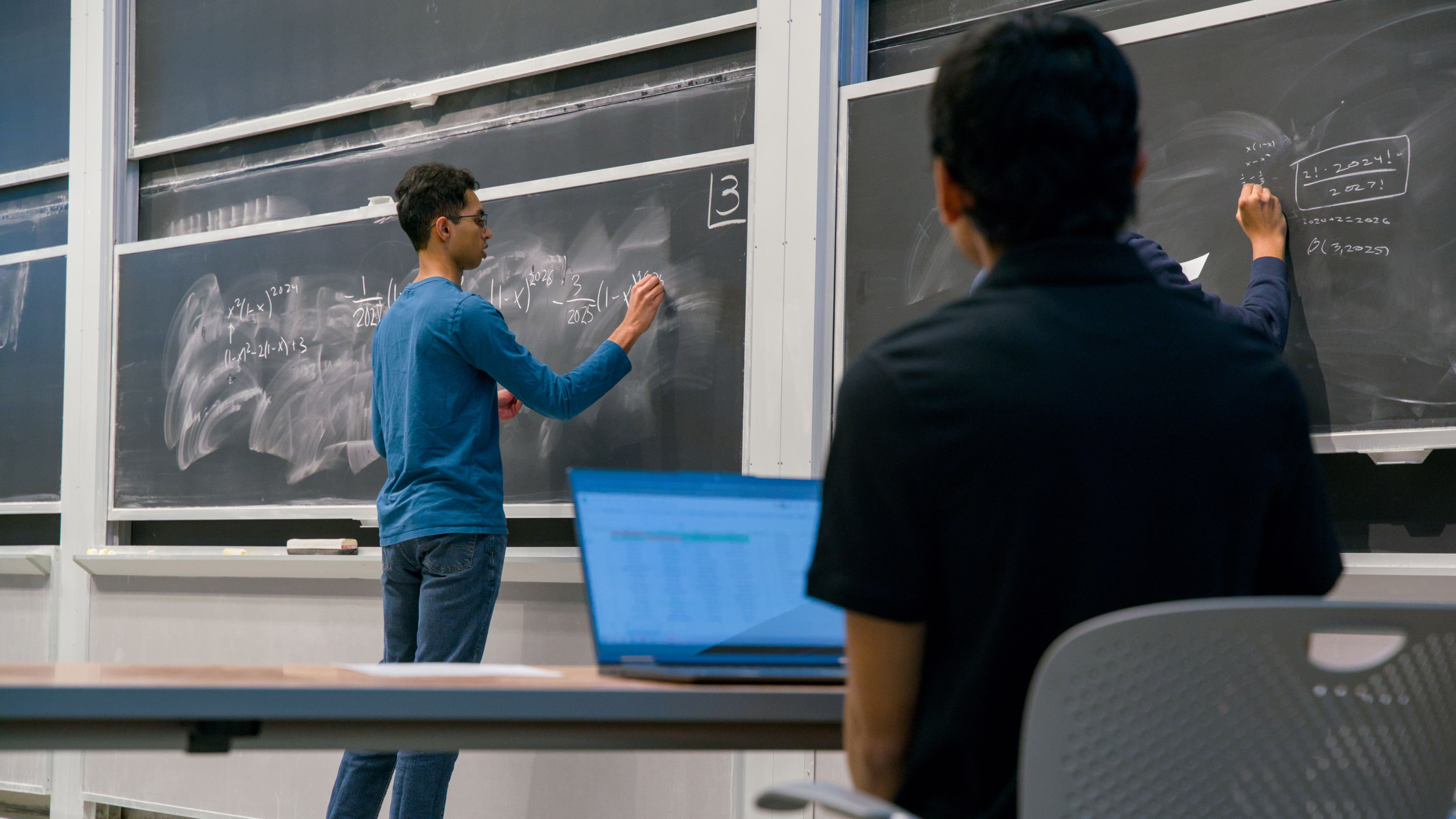 Adithya Balachandran ’25 writes on a chalkboard while someone seated in the foreground looks on.